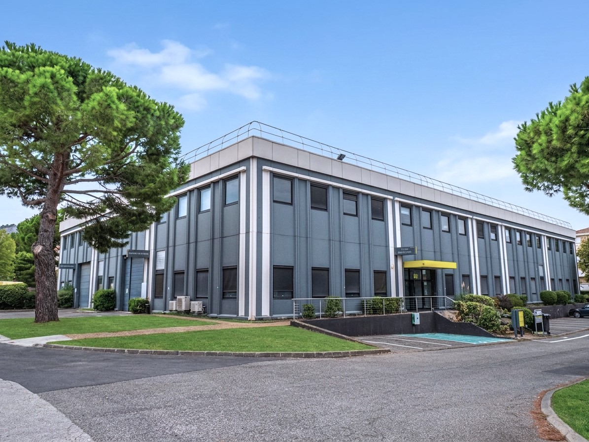 Un immeuble de bureaux moderne de deux étages, avec des panneaux gris, de grandes baies vitrées et une entrée jaune, entouré d'arbres et de pelouses verdoyantes sous un ciel bleu limpide.