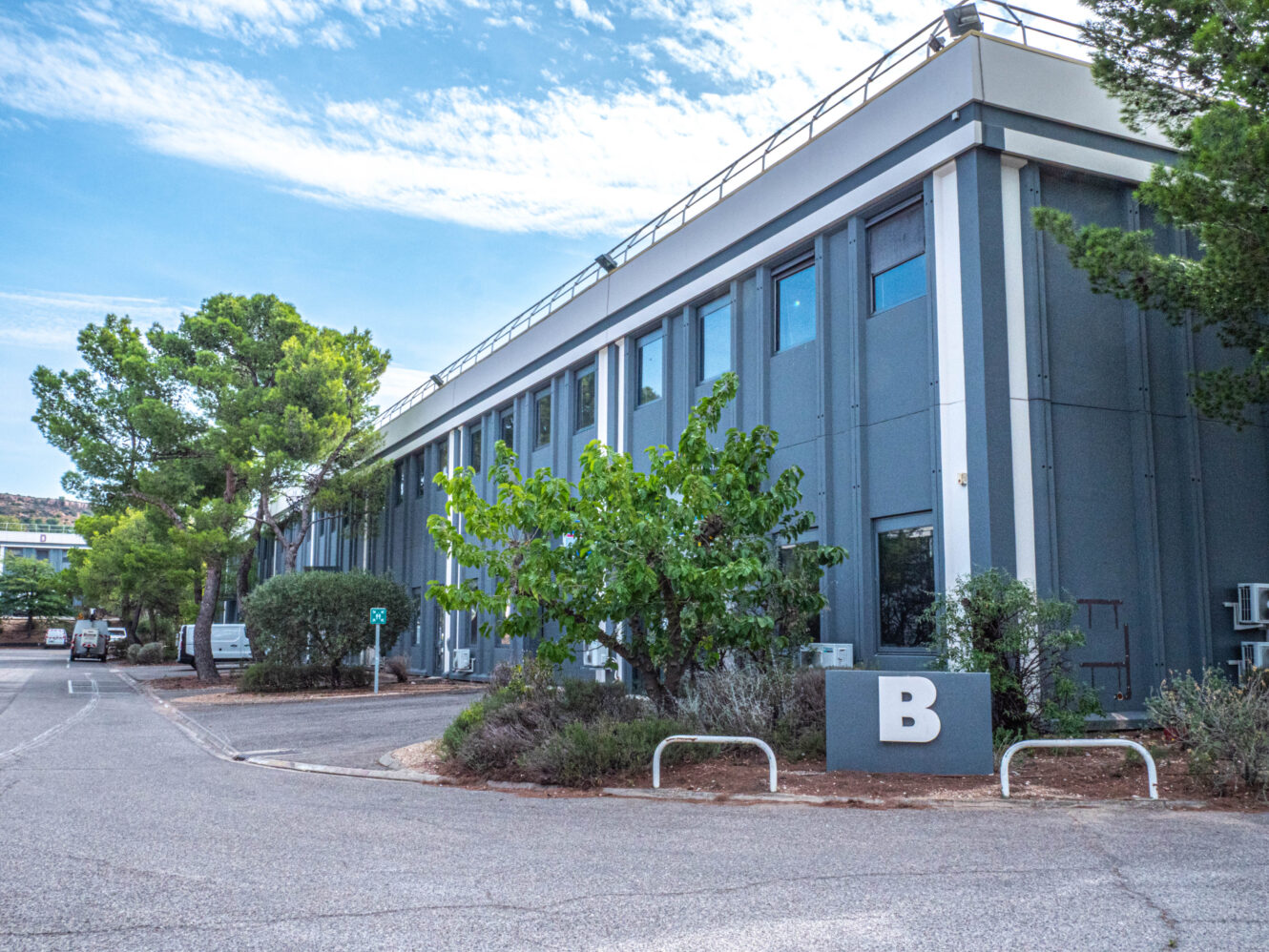 Bâtiment moderne de couleur grise, doté de nombreuses fenêtres, entouré d'arbres et d'un parking ; un grand « B » figure sur un panneau près de l'entrée.