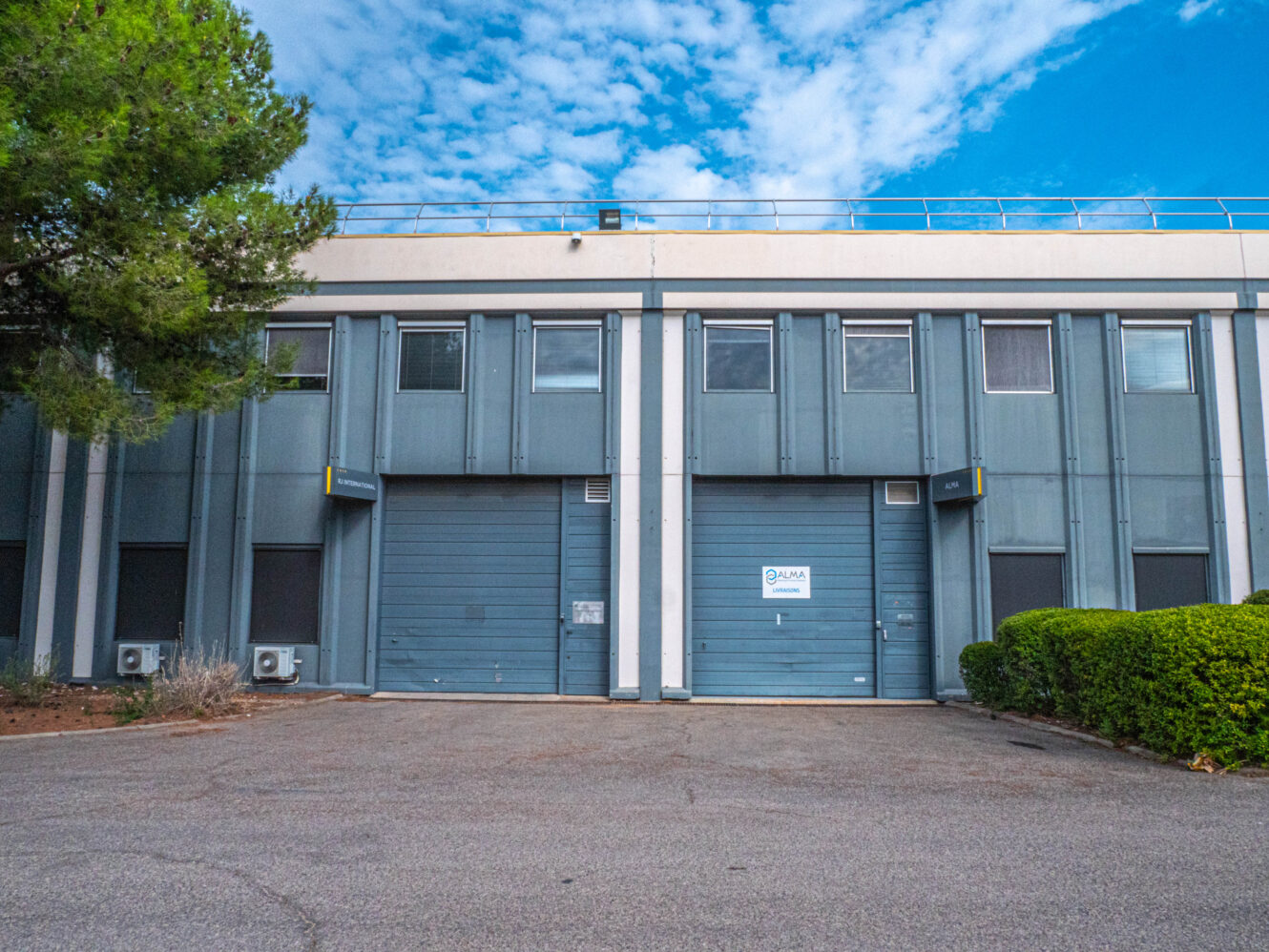 Bâtiment industriel gris doté de deux grandes portes de garage, de petites fenêtres au-dessus et d'une enseigne sur l'une des portes. On aperçoit des arbres et des buissons sur les côtés, sous un ciel partiellement nuageux.