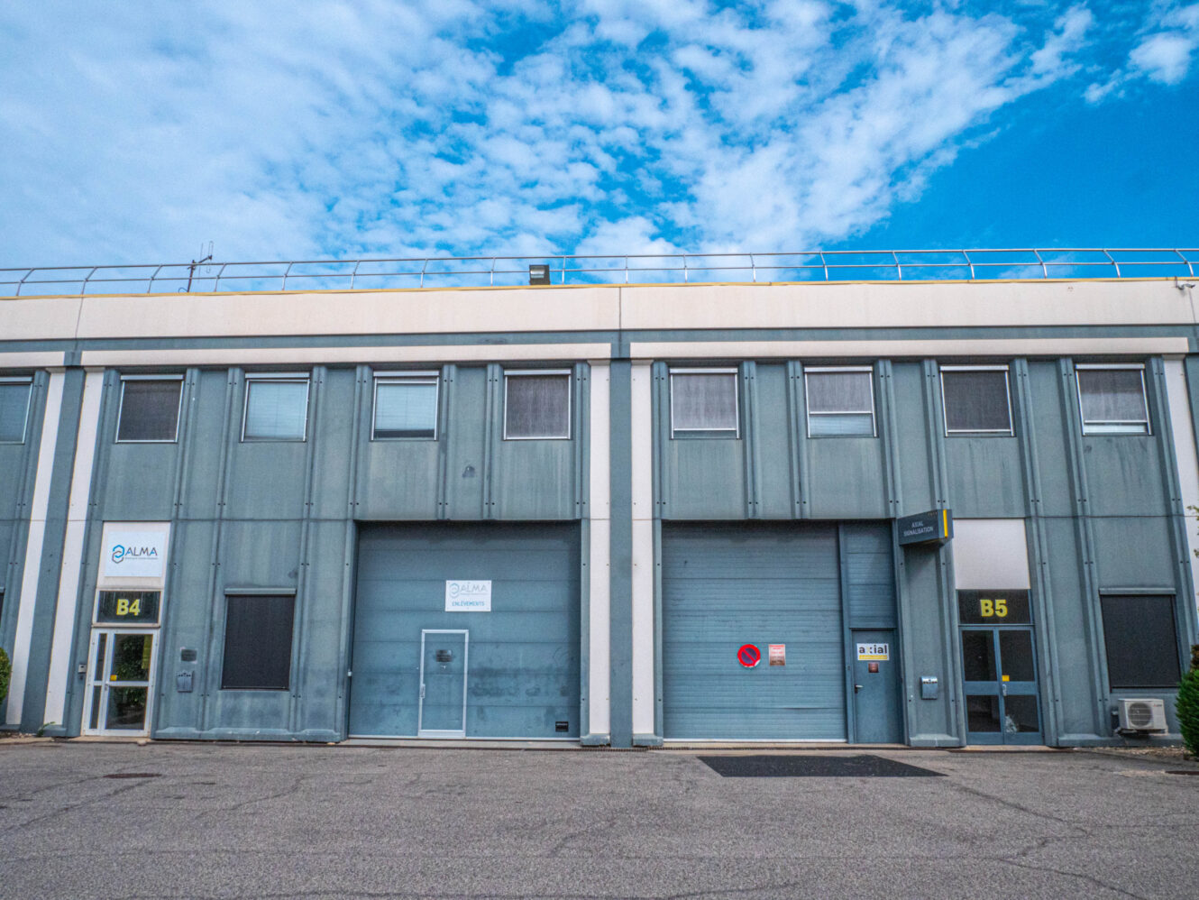 Deux bâtiments industriels, numérotés B4 et B5, dotés de portes et de fenêtres métalliques, se dressent sous un ciel bleu parsemé de nuages.