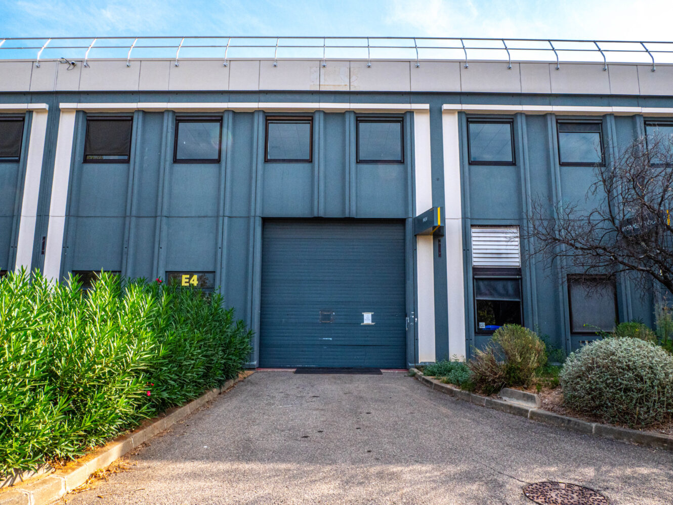 Bâtiment industriel moderne doté d'une grande porte de garage bleue, de fenêtres tout autour et d'un aménagement paysager comprenant des buissons et des arbustes au premier plan.