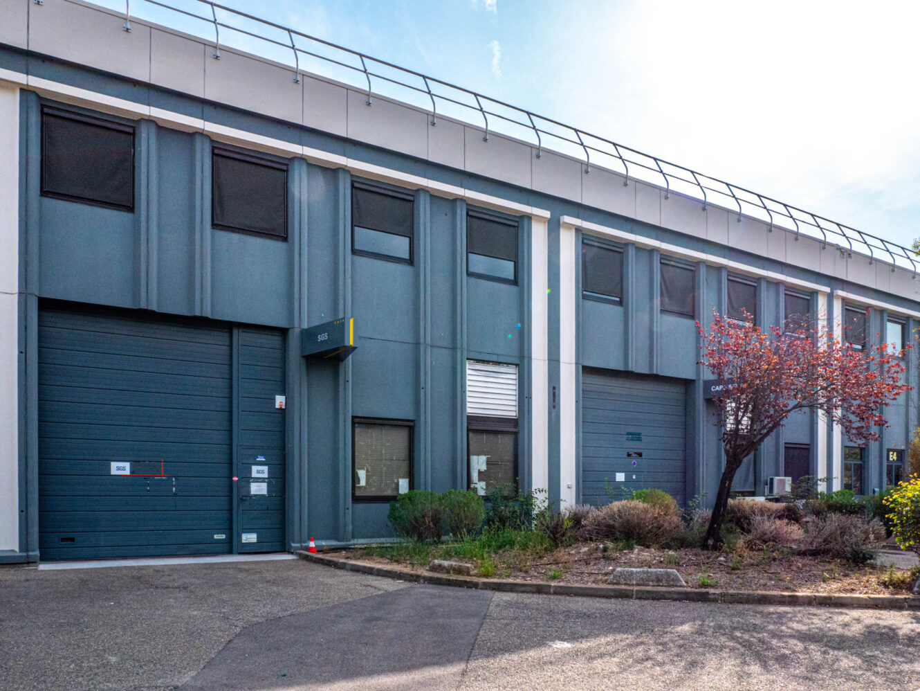 Un bâtiment industriel moderne doté de portes de garage bleu-gris, de petites fenêtres et d'un espace paysager agrémenté d'arbustes et d'un arbre à l'avant.