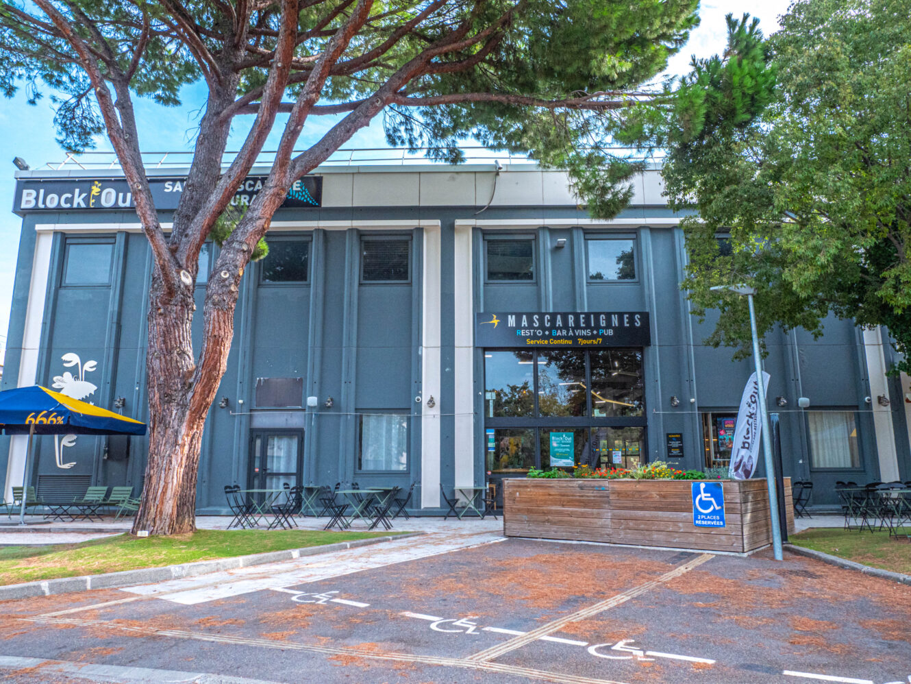Extérieur du restaurant avec terrasse, un grand pin, une place de stationnement réservée aux personnes à mobilité réduite et une signalétique bien visible pour les enseignes « Mascarignes » et « Block Out ».