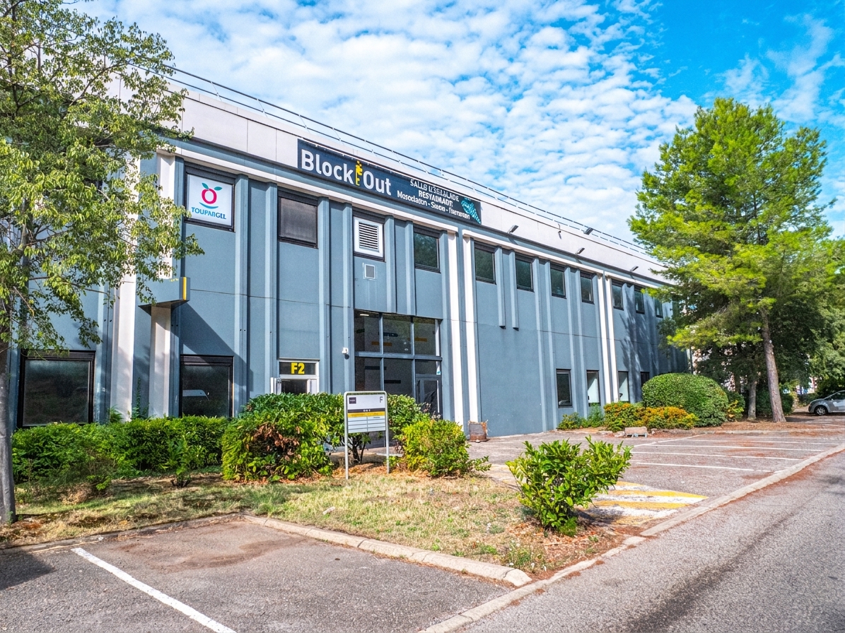 Immeuble commercial moderne de deux étages, doté d'une enseigne « BlockOut », d'un parking comportant quelques places libres, ainsi que d'arbres et d'arbustes tout autour, sous un ciel partiellement nuageux.