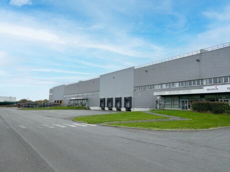 Grand entrepôt industriel doté de plusieurs quais de chargement, d'une allée pavée et d'un espace vert, sous un ciel partiellement nuageux.
