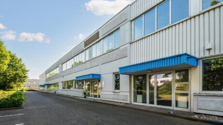 Ein modernes, zweistöckiges Bürogebäude mit großen Fenstern und blauen Markisen, das an einem klaren Tag neben einer Einfahrt steht.