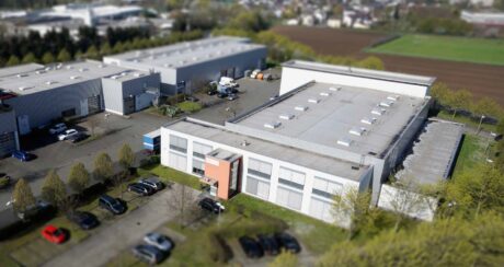 Luftaufnahme eines weißen Industriegebäudes mit einem kleinen orangefarbenen Eingang, angrenzendem Parkplatz, nahe gelegenen Lagerhäusern und grünen Feldern im Hintergrund.