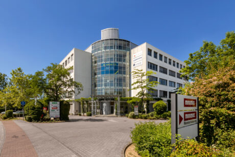 Ein modernes vierstöckiges Bürogebäude mit einem gläsernen Eingang, umgeben von Bäumen und Schildern an der Einfahrt, unter einem klaren blauen Himmel.