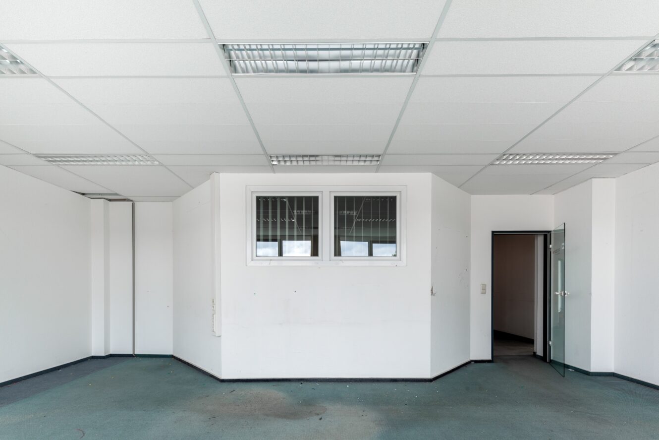 Ein leerer, weißer Büroraum mit blauem Teppich, fluoreszierenden Deckenleuchten, zwei vergitterten Fenstern und einer offenen Glastür, die in einen dunkleren Raum führt.