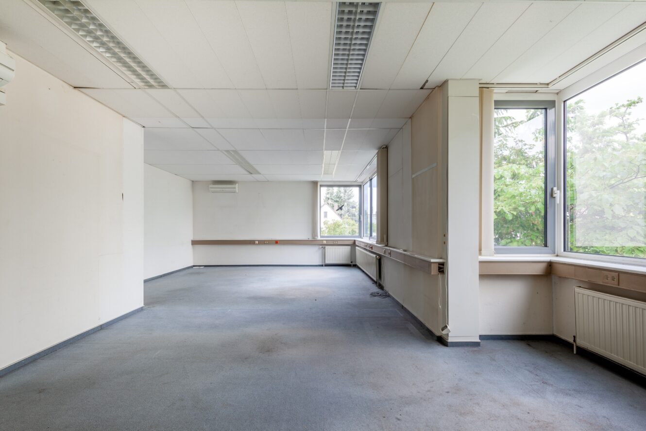 Ein leerer Büroraum mit weißen Wänden, fluoreszierenden Deckenleuchten, Teppichboden, großen Fenstern und einigen sichtbaren Steckdosen und Kabeln an den Wänden.