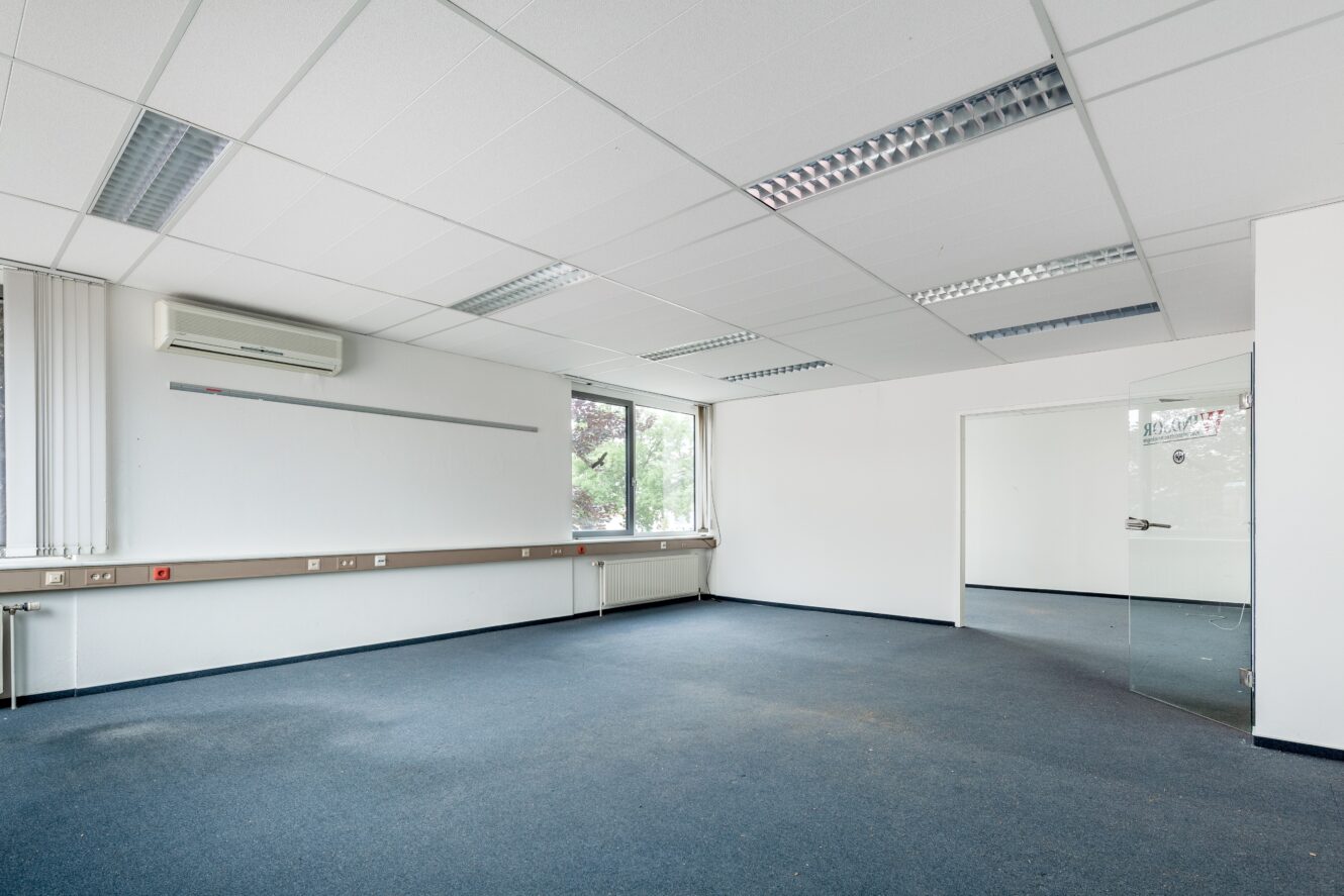 Leerer Büroraum mit blauem Teppich, weißen Wänden, fluoreszierenden Deckenleuchten, großen Fenstern, Glastür und einer Klimaanlage an der Wand.