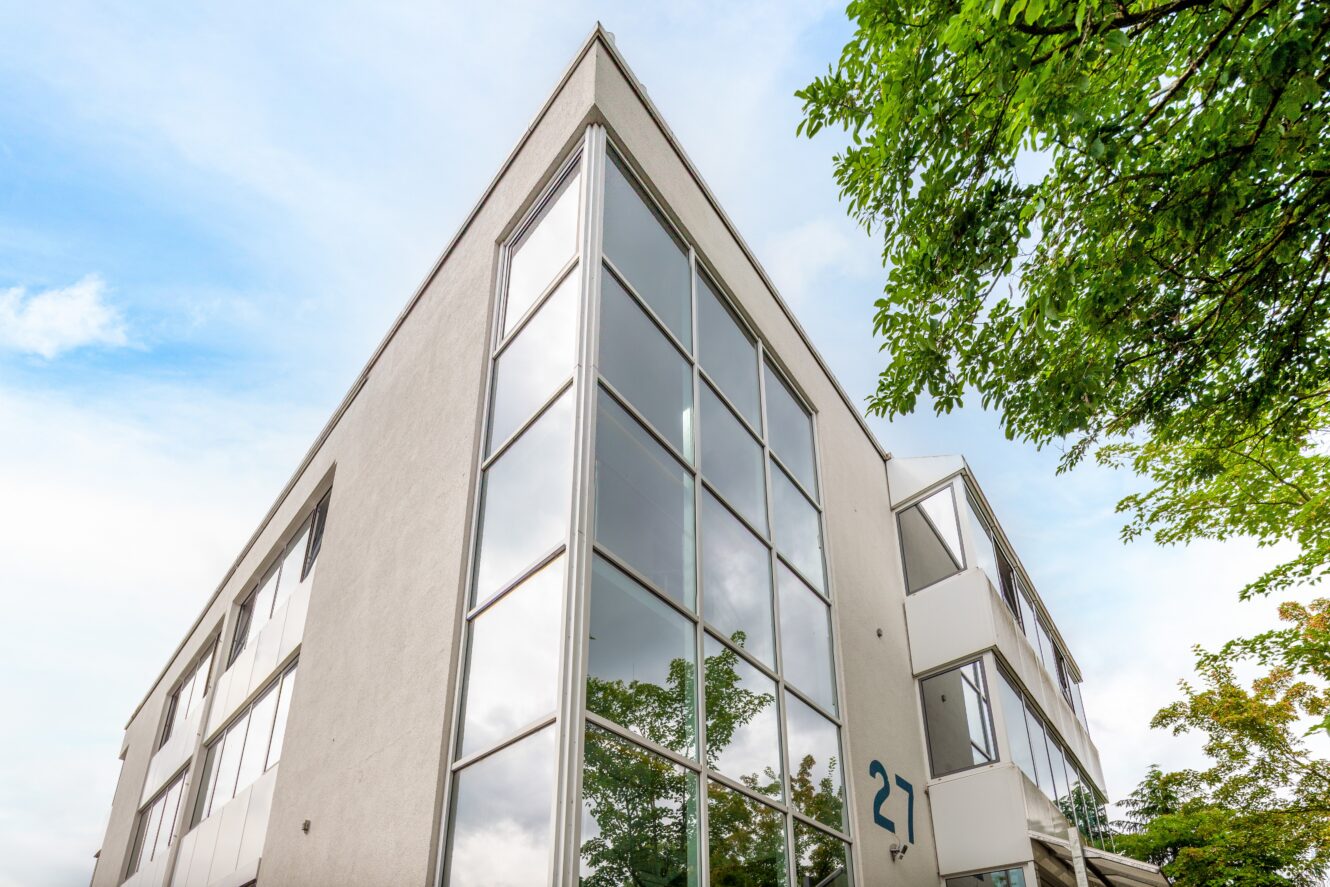 Ein modernes dreistöckiges Gebäude mit großen Glasfenstern und der Zahl 21 in der Nähe des Eingangs; Bäume und Himmel spiegeln sich in den Fenstern.