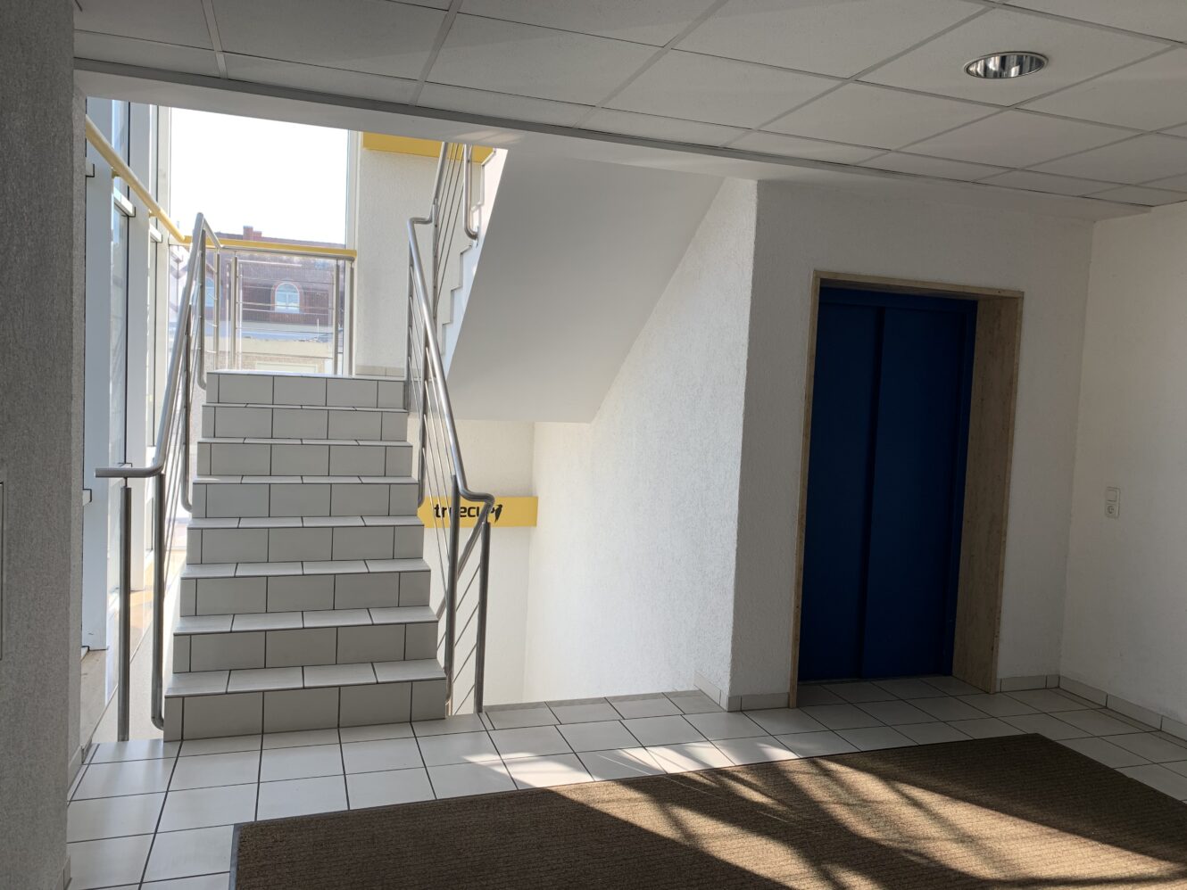 Ein gefliester Flur mit einer Treppe auf der linken Seite, einer blauen Aufzugstür auf der rechten Seite und Sonnenlicht, das durch die Fenster fällt.