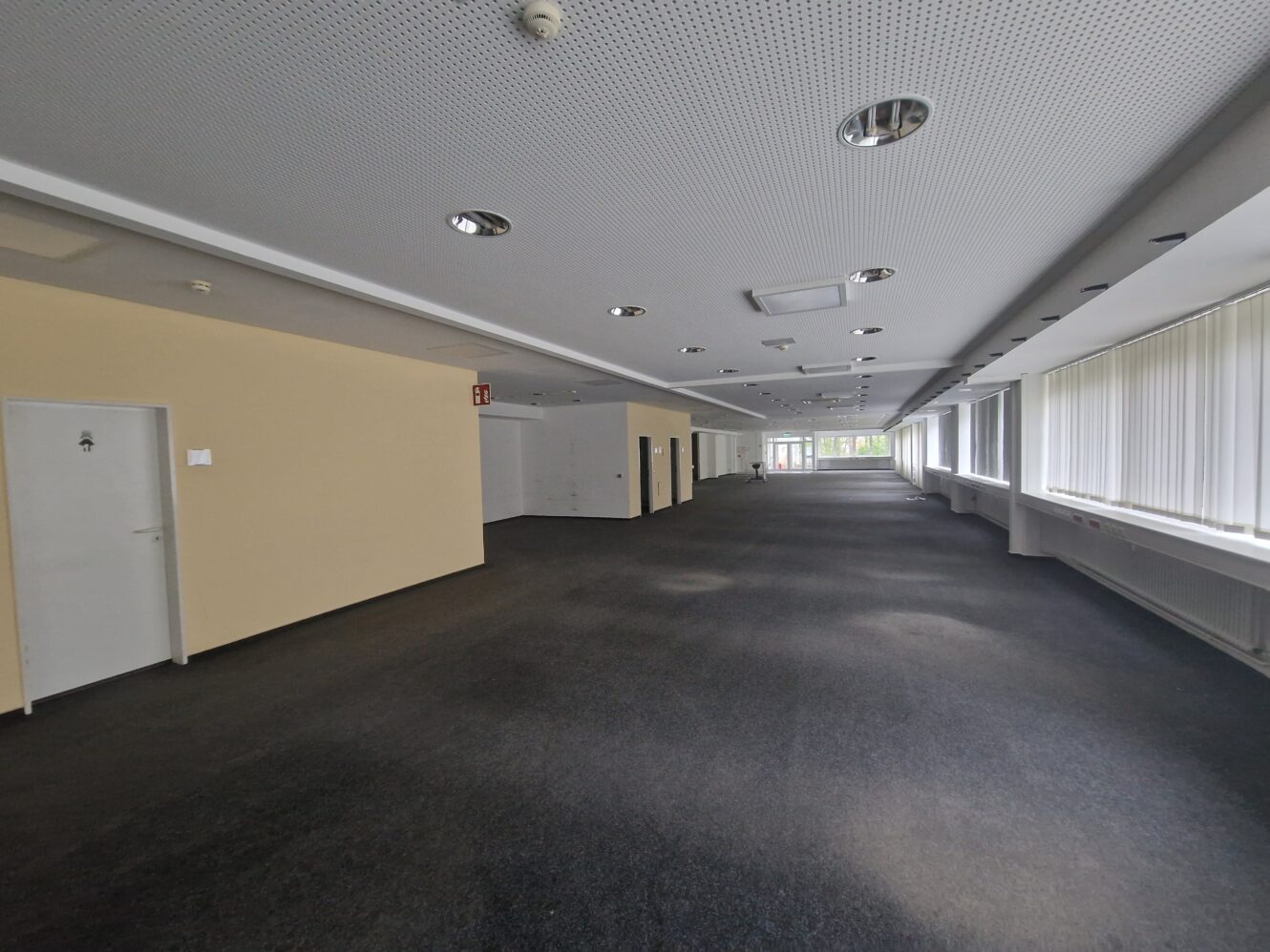 Ein geräumiger, leerer Büroflur mit Teppichboden, weißen Wänden und großen Fenstern auf der rechten Seite, die viel Tageslicht hereinlassen.