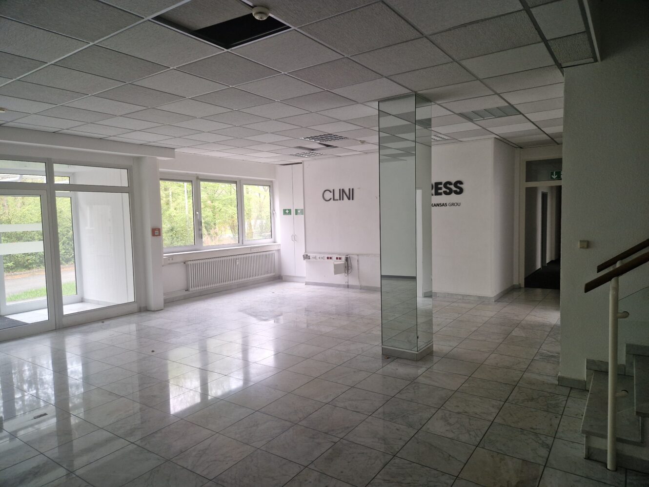Leerer Raum mit Marmorböden, großen Fenstern, Glastüren und weißen Wänden; an der Wand befinden sich teilweise Schilder mit der Aufschrift CLINI und ESS.