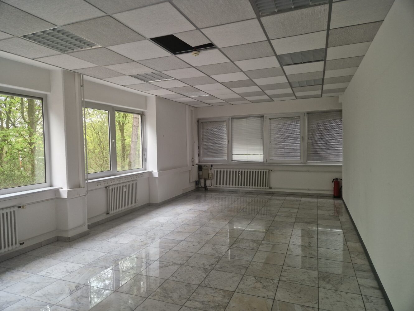 Leerer Büroraum mit gefliestem Boden, weißen Wänden, mehreren Fenstern und einigen fehlenden oder deplatzierten Deckenplatten. An der Rückwand sind ein Feuerlöscher und ein Heizkörper zu sehen.