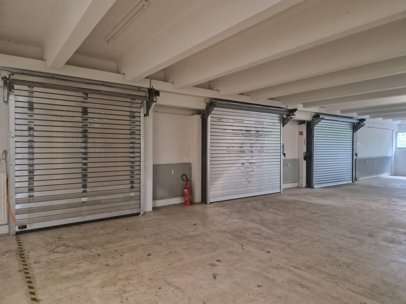 Drei metallene Garagenrolltore in einem leeren, sauberen Parkplatz mit Betonboden, weißen Wänden und einem Feuerlöscher zwischen den ersten beiden Toren.