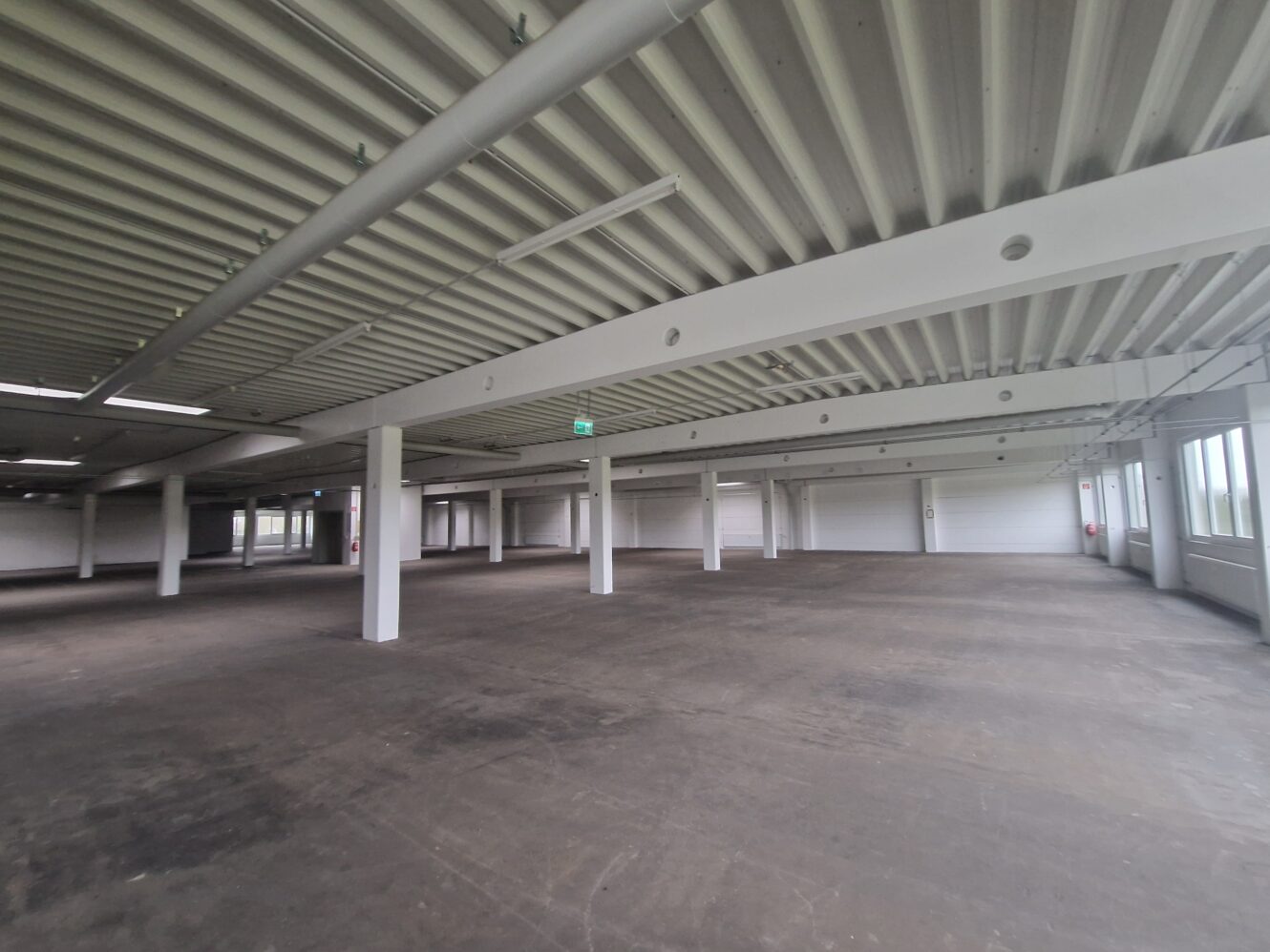 Geräumiges, leeres Industrielager mit Betonboden, weißen Säulen und Leuchtstoffröhrenbeleuchtung. Große Fenster und Notausgangsschild sichtbar.