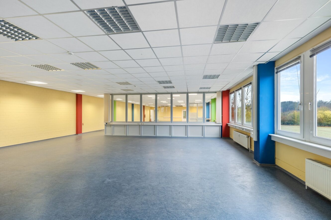 Leerer, hell erleuchteter Raum mit großen Fenstern, blauen und gelben Wänden, gekachelter Decke und einer Glastrennwand im Inneren mit einem Tresen.