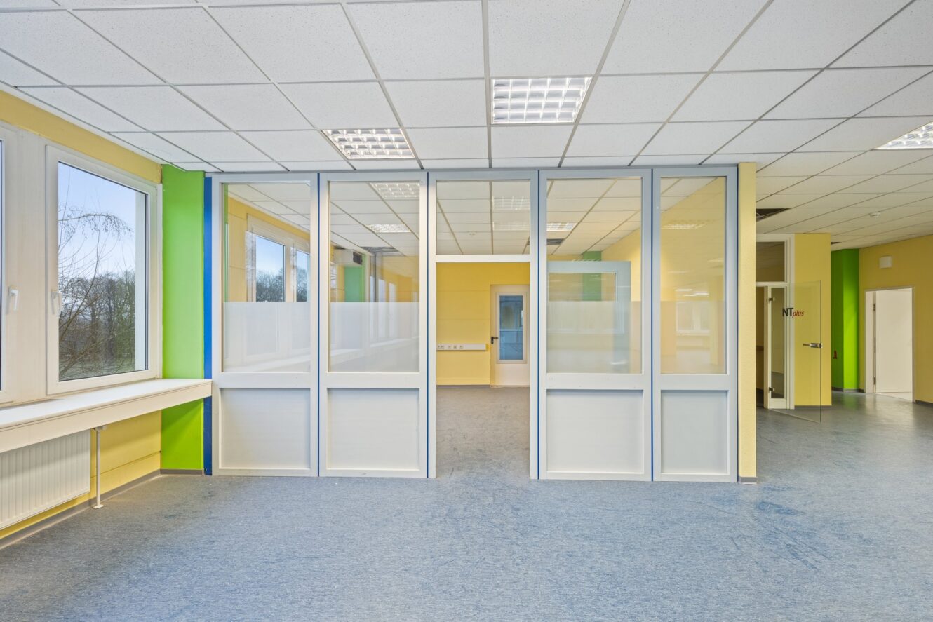 Ein hell erleuchteter, leerer Büroraum mit blauem Fußboden, grünen und gelben Wänden, großen Fenstern und einer Glastrennwand mit einer Tür, die den Raum teilt.
