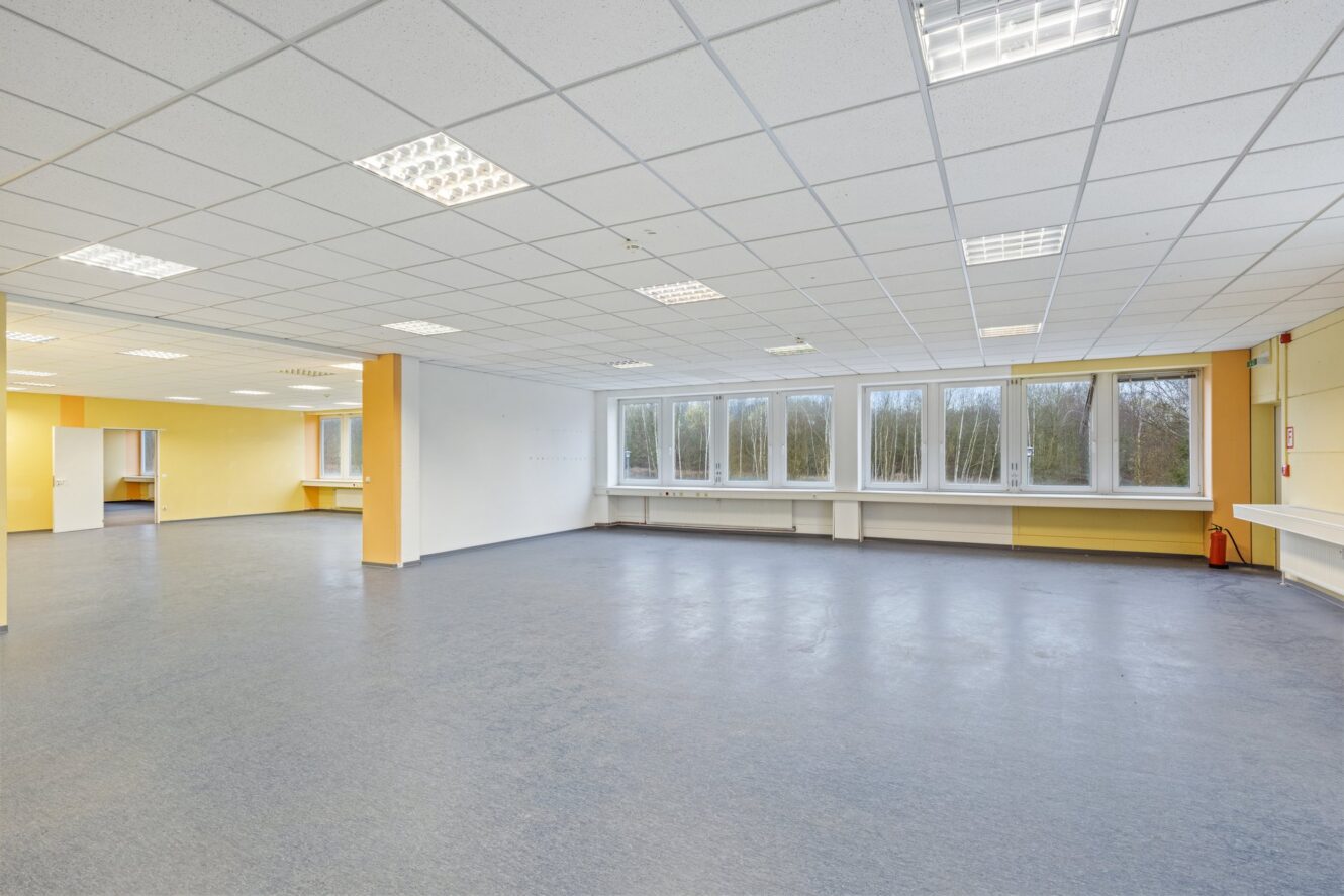 Ein großer, leerer Raum mit blauem Fußboden, weißen Deckenfliesen, Leuchtstoffröhren und mehreren Fenstern an einer Wand. Die Wände sind weiß und gelb gestrichen.