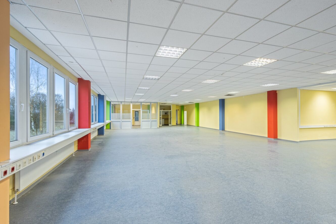 Ein großer, leerer, hell erleuchteter Raum mit einer gekachelten Decke, großen Fenstern auf einer Seite und bunten Säulen an hellgelben Wänden.