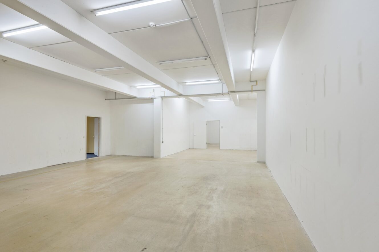 Leerer, gut beleuchteter Industrieraum mit weißen Wänden, freiliegenden Deckenbalken und einem beigen Betonboden; an den Wänden sind einige Türen zu sehen.
