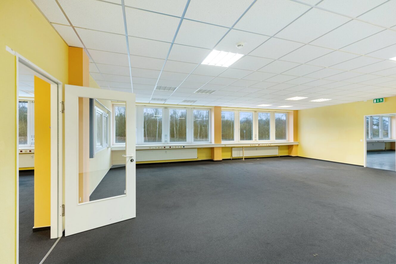 Großer leerer Raum mit gelben Wänden, grauem Teppich, einer weißen Decke mit Leuchtstoffröhren, mehreren Fenstern und einer offenen Tür, die zu einem anderen ähnlichen Raum führt.