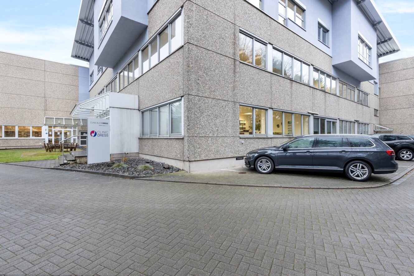 Ein modernes Bürogebäude mit großen Fenstern, ein geparkter schwarzer Kombi und ein Schild in der Nähe des Eingangs auf einer gepflasterten Zufahrt.