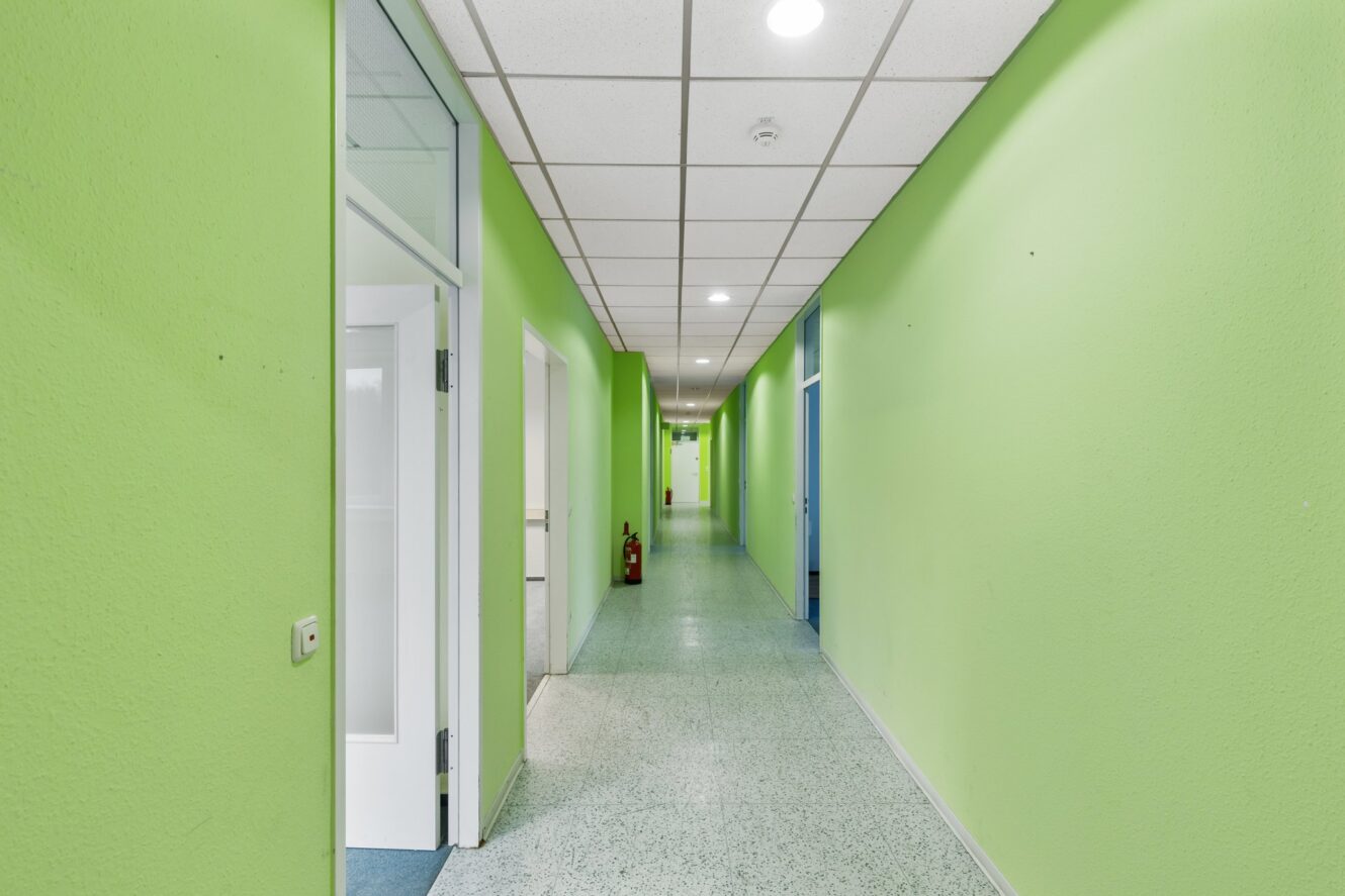 Ein langer Flur mit hellgrünen Wänden, einer weißen Decke mit Einbaulampen und mehreren offenen Türen, die zu angrenzenden Räumen führen. In der Ferne ist ein Feuerlöscher zu sehen.