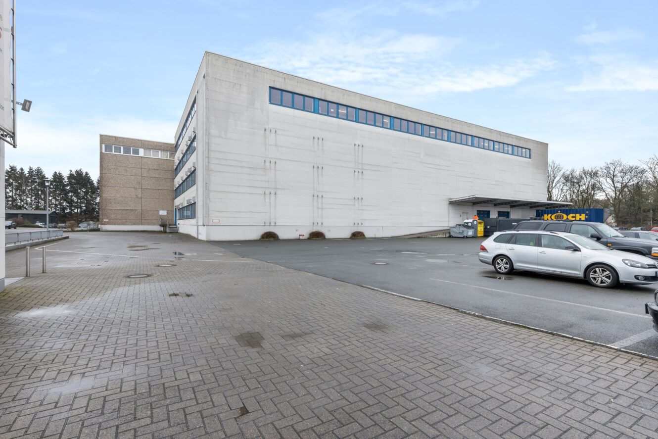 Ein großes, weißes Industriegebäude mit vielen Fenstern und einem Parkplatz mit mehreren Autos an einem bewölkten Tag.