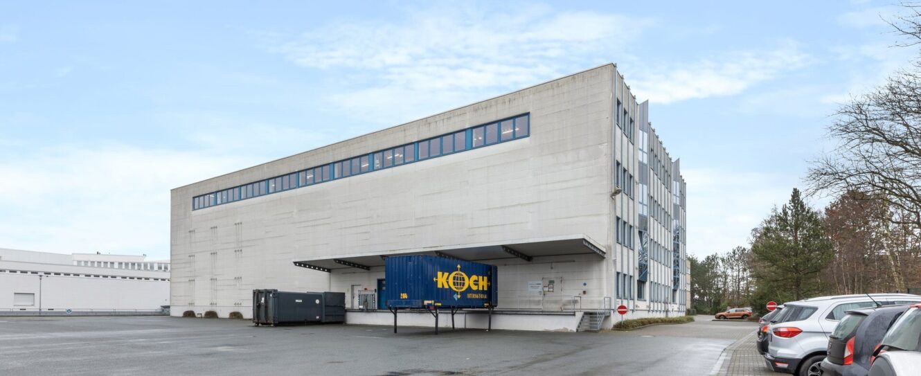 Ein großes, weißes Industriegebäude mit einer Laderampe und einem blauen Frachtcontainer mit der Aufschrift KOCH neben geparkten Autos unter einem bewölkten Himmel.