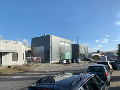 Moderne Industriebauten mit großen Fenstern und flachen Dächern entlang einer von parkenden Autos gesäumten Straße unter einem klaren blauen Himmel.