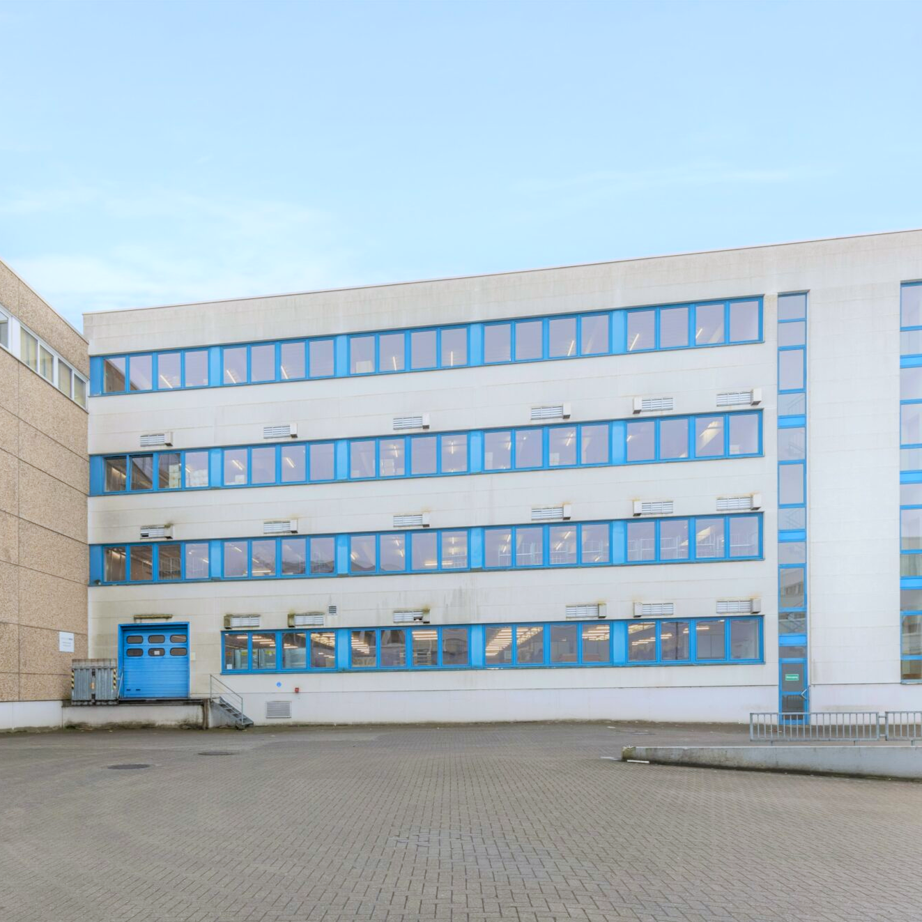 Ein vierstöckiges weißes Gebäude mit blauen Fensterrahmen und Türen, das neben einer leeren, gepflasterten Fläche unter freiem Himmel steht.