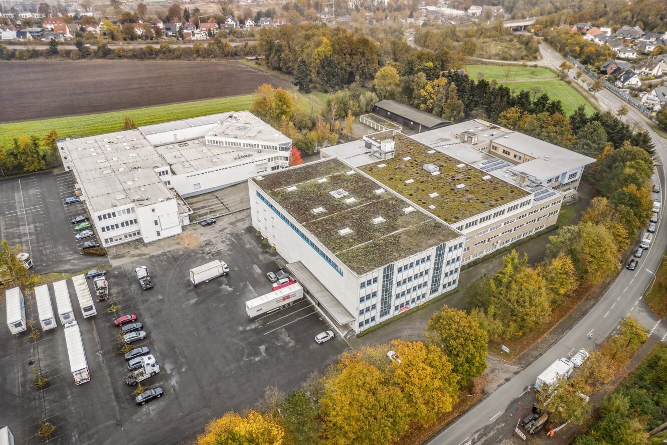 Luftaufnahme eines Industriekomplexes mit mehreren großen Gebäuden, einem Parkplatz mit Autos und Lastwagen und umliegenden Bäumen und Feldern in einer herbstlichen Umgebung.