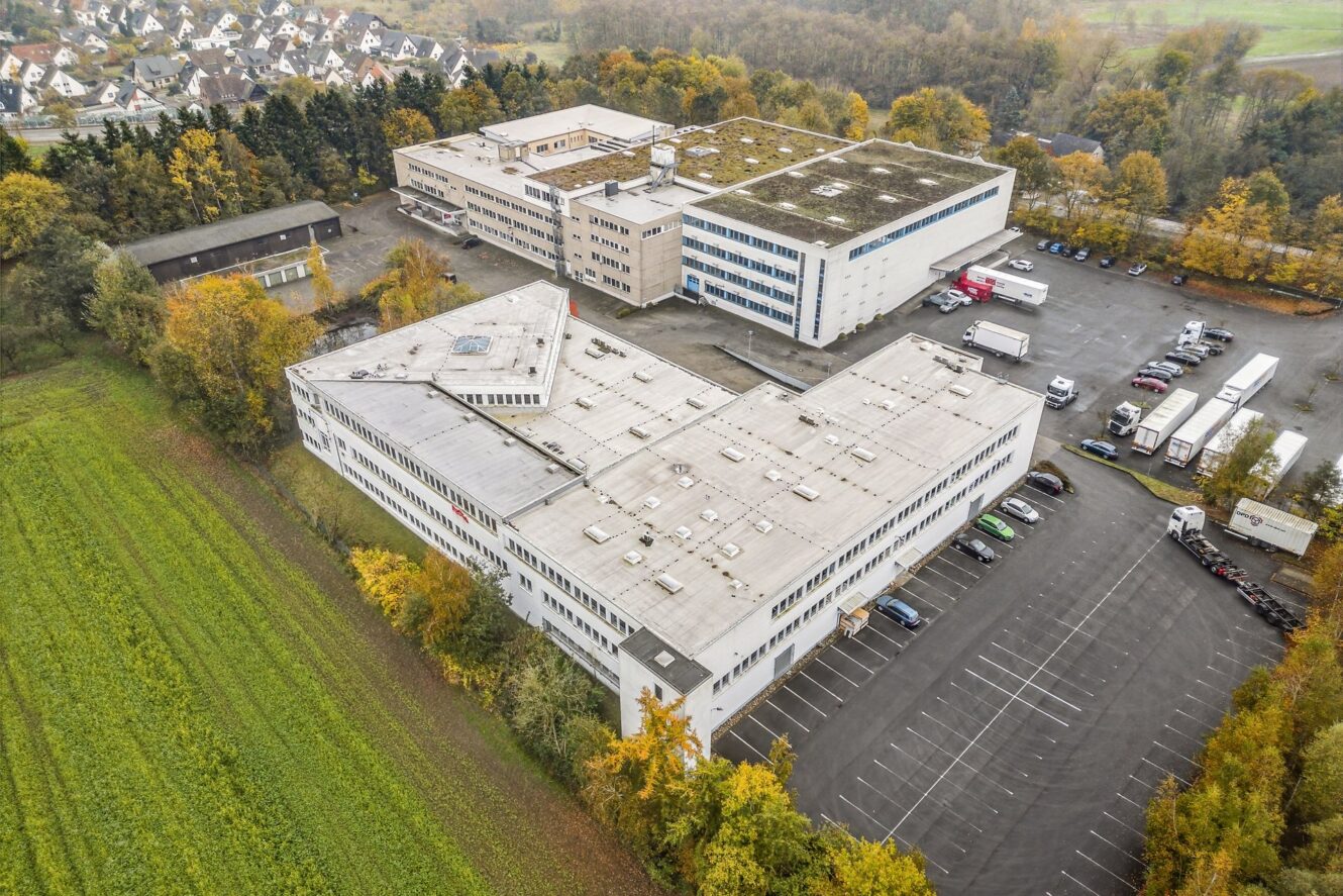 Luftaufnahme eines großen Industrie- oder Bürokomplexes mit mehreren Gebäuden, Parkplätzen und Lastwagen, umgeben von Bäumen und Feldern in einer herbstlichen Umgebung.