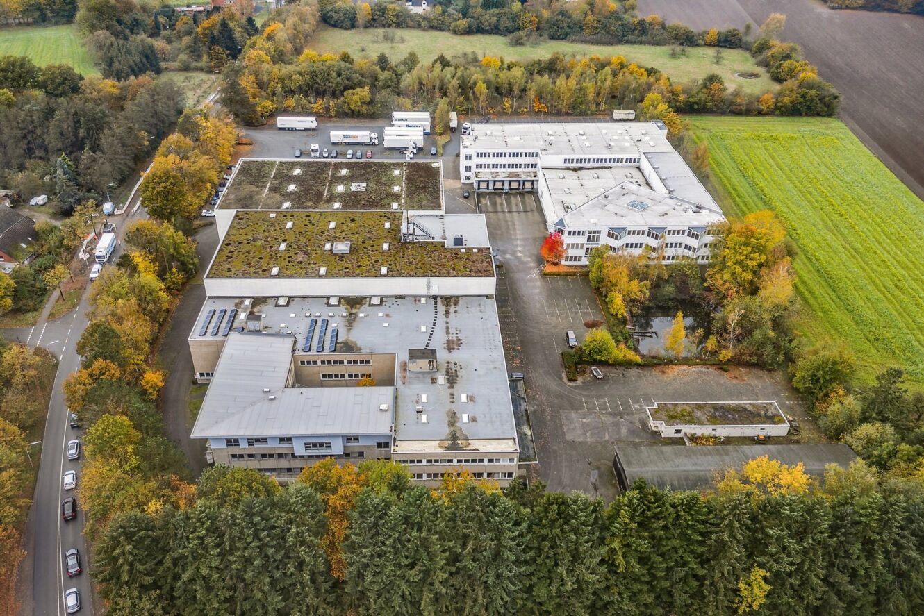 Luftaufnahme eines großen Industrie- oder Bürokomplexes, umgeben von Bäumen, mit Parkplätzen, geparkten Fahrzeugen und angrenzenden Feldern im Herbst.