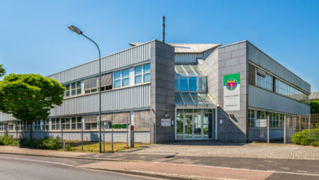 Modernes zweistöckiges Bürogebäude mit großen Fenstern, einem auffälligen Schild in der Nähe des Eingangs und einem Baum auf dem Gehweg an einem klaren, sonnigen Tag.