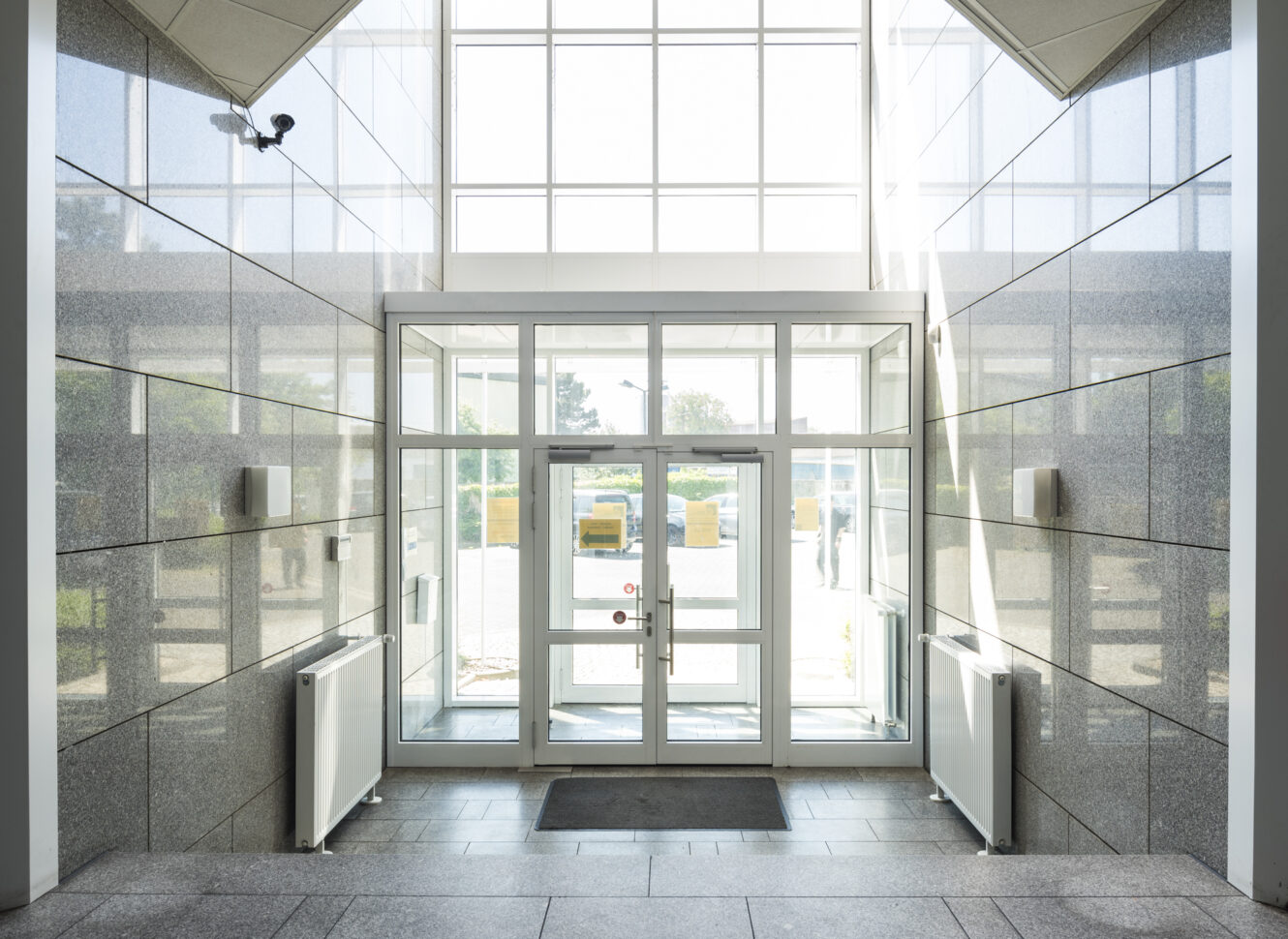 Moderner Gebäudeeingang mit Glastüren, großen Fenstern, gefliesten Wänden und Heizkörpern auf beiden Seiten, die zu einem durch das Glas sichtbaren Außenbereich führen.
