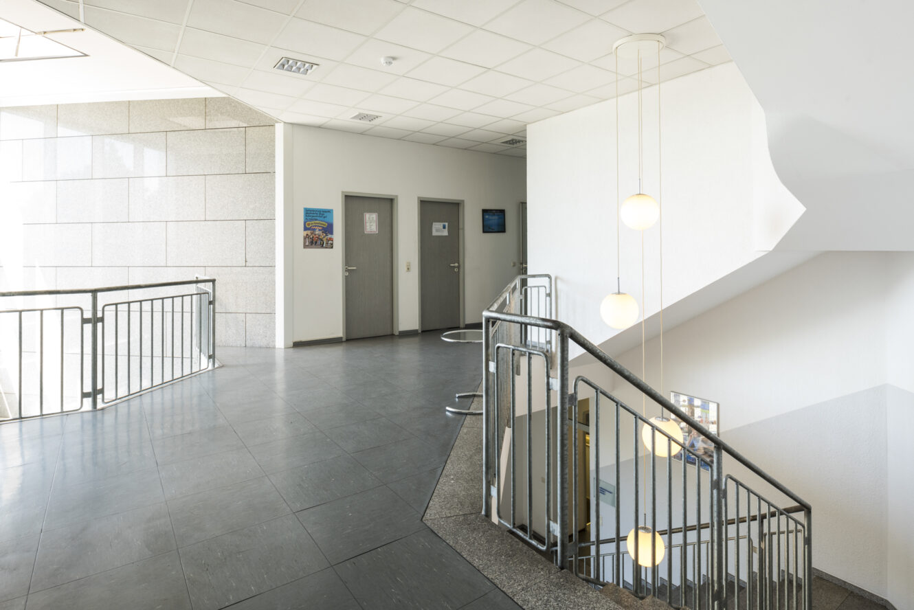 Innenansicht eines modernen Gebäudeflurs mit drei geschlossenen Türen, einer Treppe mit Metallgeländer und hängenden Kugellampen.