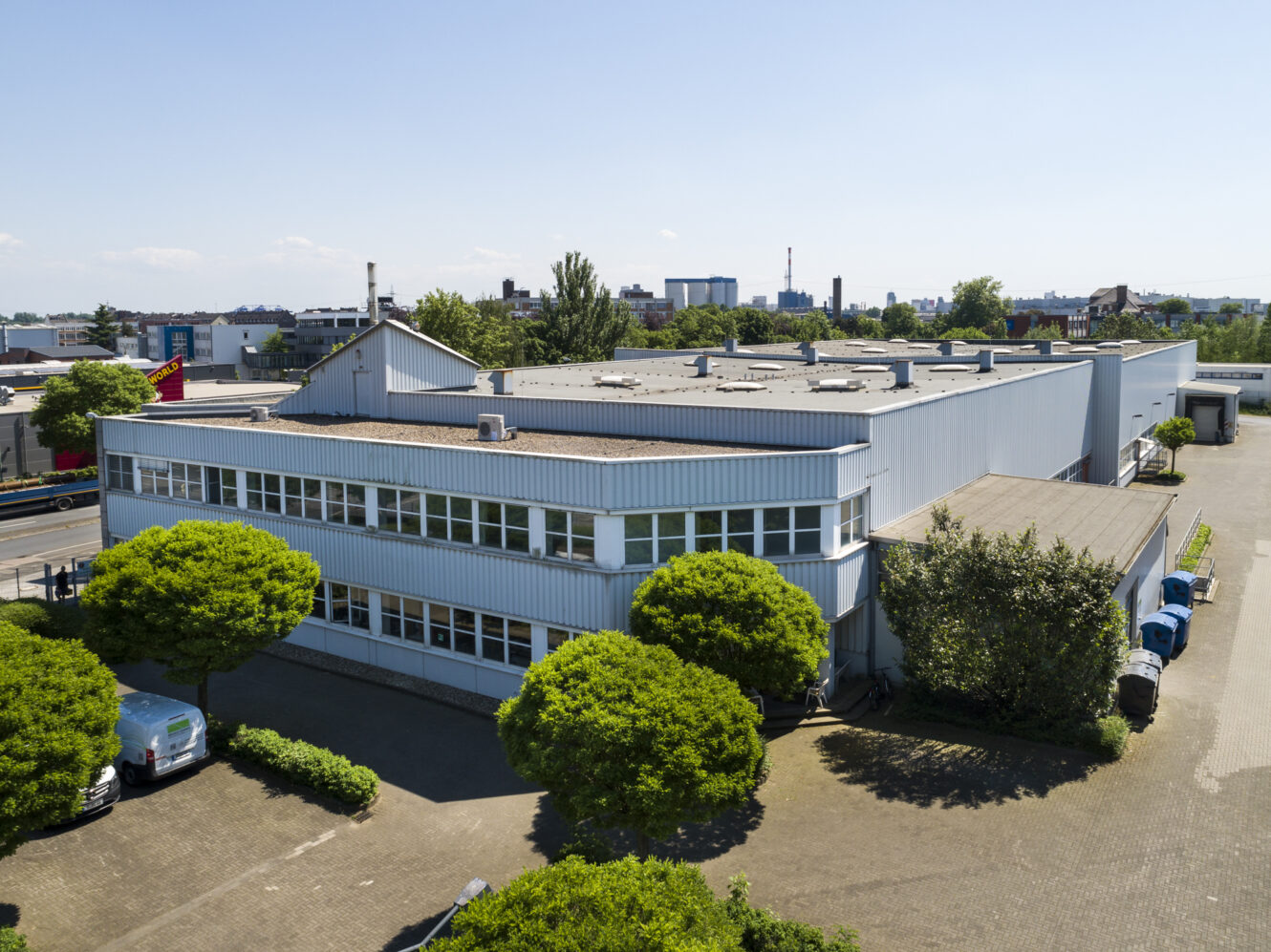 Ein großes hellblaues Industrielager, umgeben von Bäumen und geparkten Fahrzeugen, unter einem klaren Himmel.