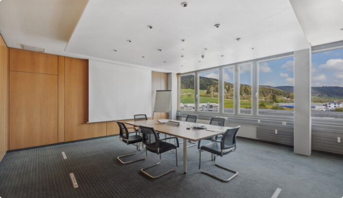 Ein moderner Konferenzraum mit einem rechteckigen Tisch, sechs Stühlen, einer Projektionsfläche, einem Flipchart und großen Fenstern mit Blick auf eine malerische Landschaft.