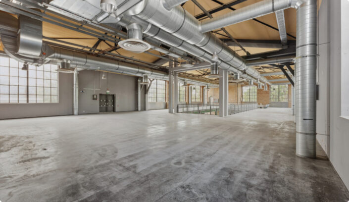 Großer leerer Industrieraum mit freiliegenden Schächten, hohen Decken, Betonböden und großen Gitterfenstern, die viel natürliches Licht hereinlassen.