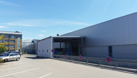 Außenansicht eines großen Industrielagers mit angrenzendem Parkplatz und Parkverbotsschildern bei strahlend blauem Himmel.