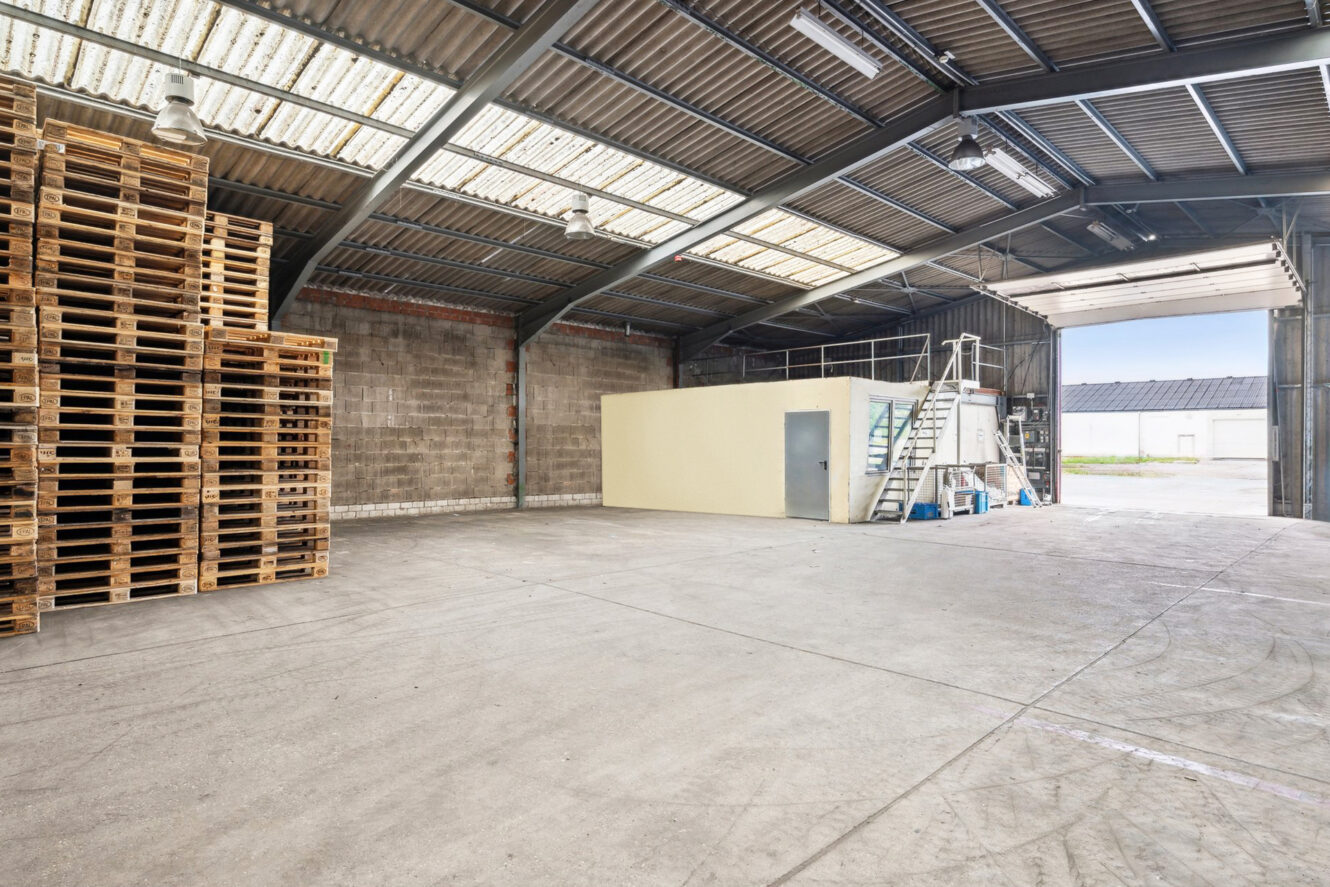 Das Innere eines leeren Lagerhauses mit einem Stapel Holzpaletten auf der linken Seite, einem kleinen geschlossenen Raum mit einer Metalltreppe und großen offenen Türen, die Tageslicht hereinlassen.