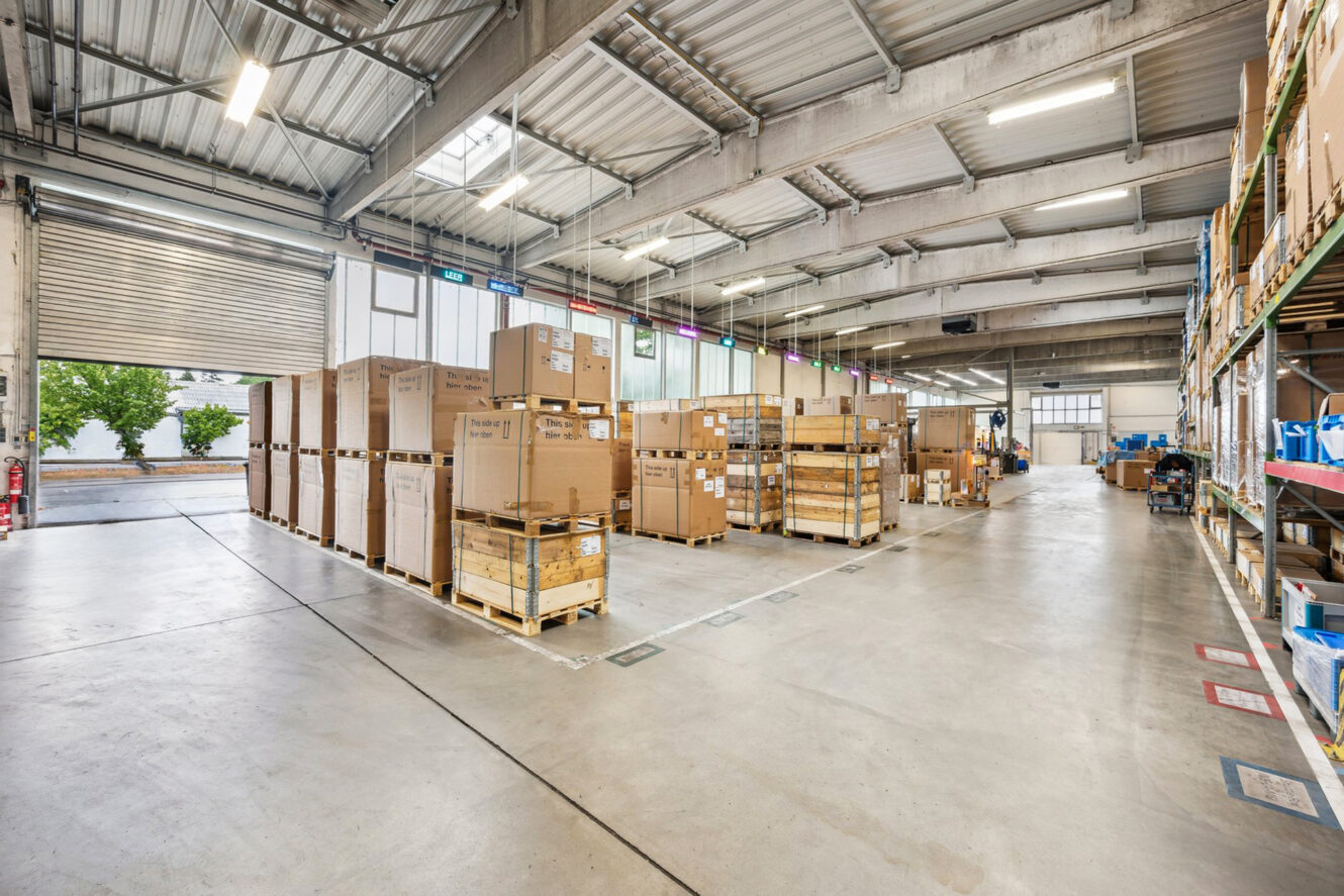Ein geräumiges Lagerhaus, in dem große Kartons und Holzpaletten ordentlich gestapelt sind, Betonböden, hohe Decken und offene Ladetore.