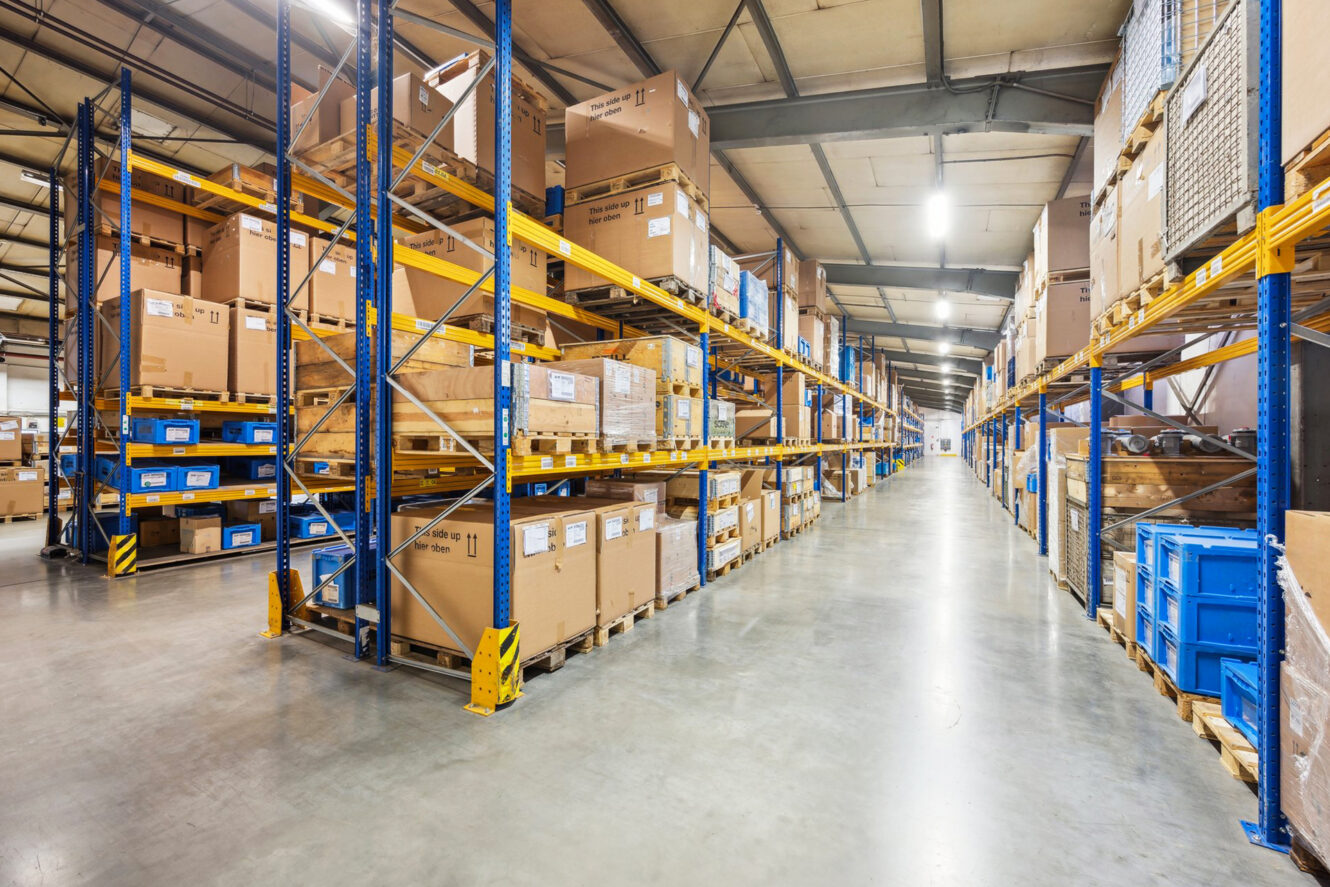 Breiter Gang zwischen hohen Lagerregalen, die mit Kartons und Lagerbehältern gefüllt sind, unter heller Deckenbeleuchtung.