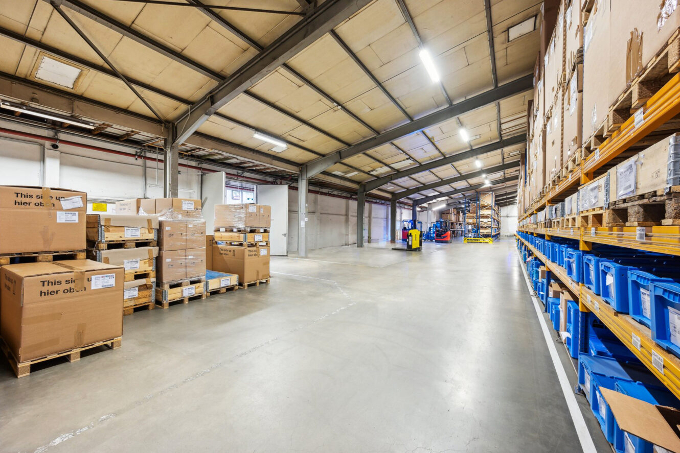 Weite Ansicht eines Lagerhauses mit gestapelten Kartons auf Paletten auf der linken Seite und blauen Kunststoffbehältern auf Regalen auf der rechten Seite, unter heller Deckenbeleuchtung.