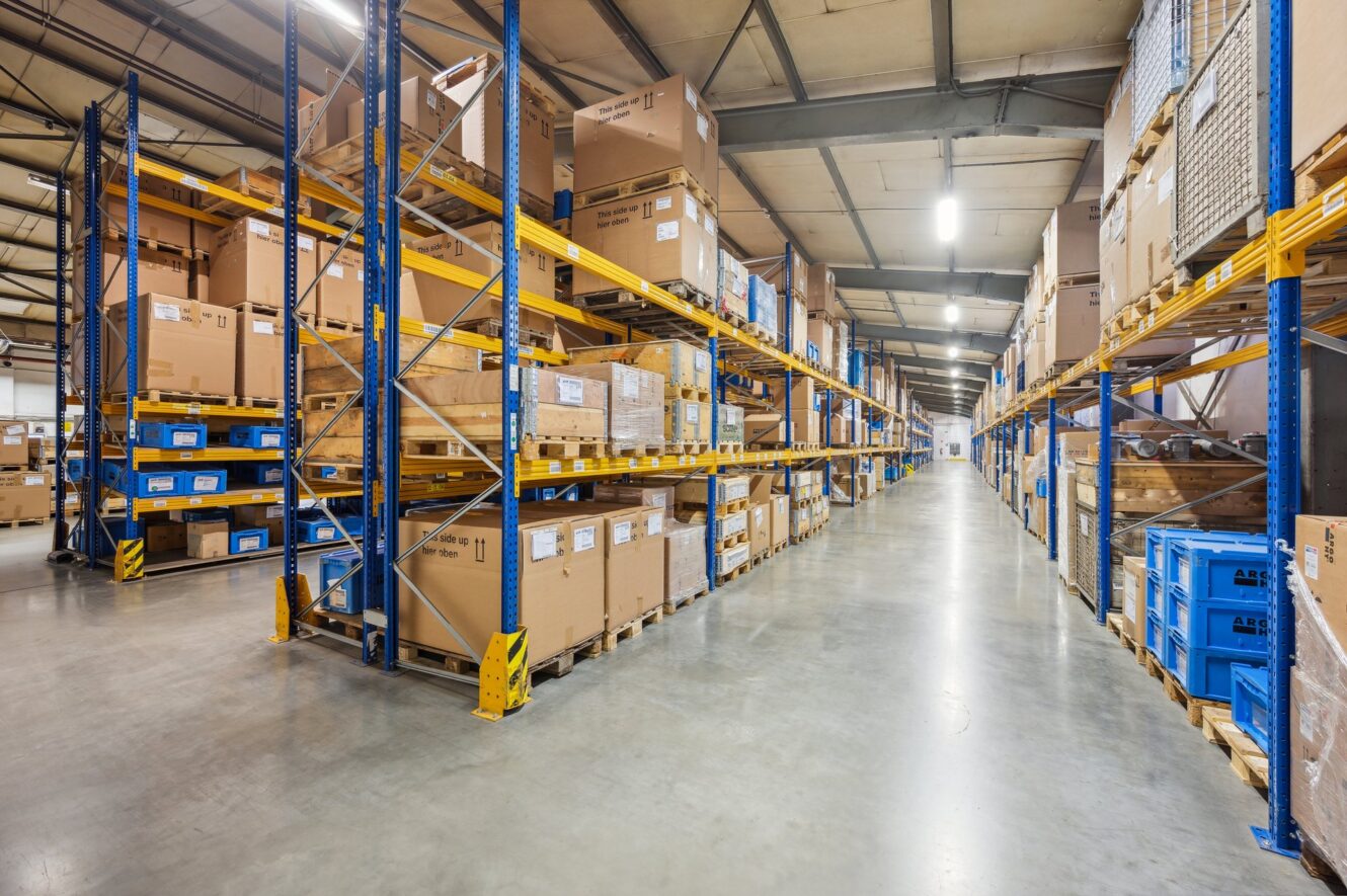 Ein breiter Gang zwischen hohen Lagerregalen, die mit großen Kartons und Plastikbehältern gefüllt sind, unter hellen Neonröhren an der Decke.