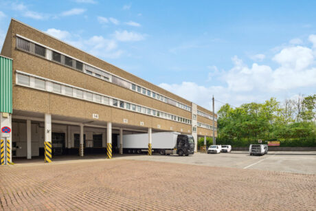 Ein großes Industriegebäude mit mehreren Laderampen; ein Sattelschlepper steht rückwärts an einer Rampe, und zwei Lieferwagen sind in der Nähe unter einem teilweise bewölkten Himmel geparkt.